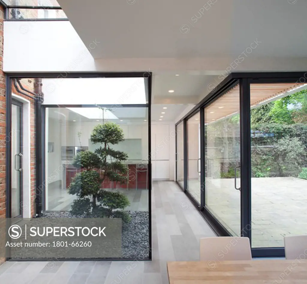 H2 Architecture, Crockerton Road, Sw17, Kitchen Extension Living Space With Glass Frontage And Void. Angle Along Glass Doors Looking Though To  Kitchen Counter  With Tree In The Mid Ground And Dinning Table In Foreground Showing Section Of Glass In Skylight Doors And Void