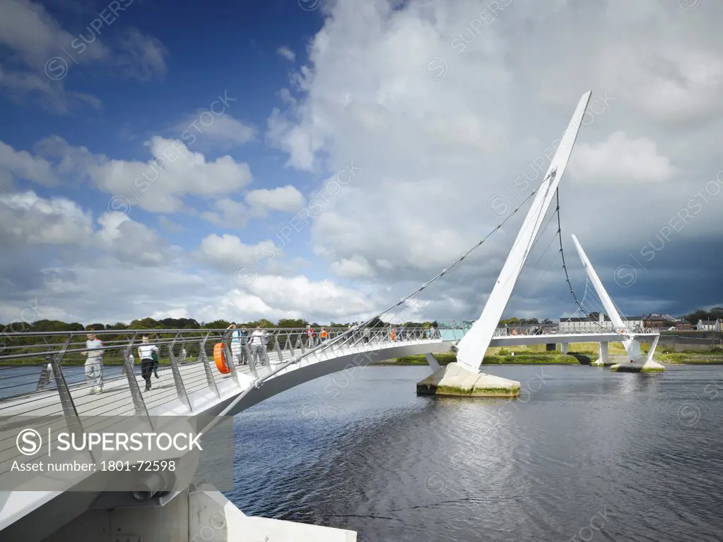 Peace Bridge, Derry, United Kingdom. Architect Wilkinson Eyre Architects, 2011. View from riverbank showing bridge with river.