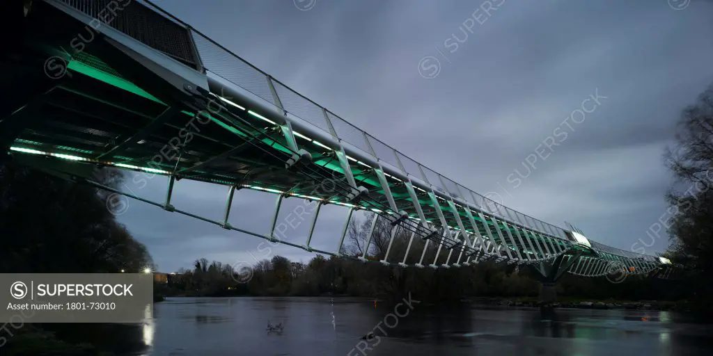 University of Limerick: Living Bridge, Limerick, Ireland. Architect Wilkinson Eyre Architects, 2007. View of from riverbank showing underside of bridge, river and surrounding landscape at dusk.
