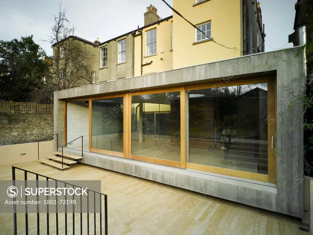 Jigsaw House, Dublin, Ireland. Architect McCullough Mulvin, 2012. View of extension showing existing house, timber decking, exposed concrete walls and sliding doors with door opened.