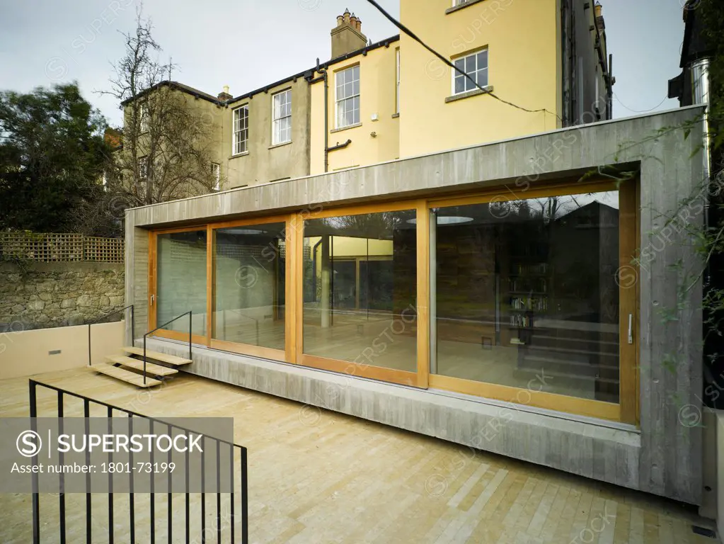 Jigsaw House, Dublin, Ireland. Architect McCullough Mulvin, 2012. View of extension showing existing house, timber decking, exposed concrete walls and sliding doors.