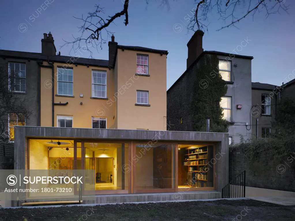 Jigsaw House, Dublin, Ireland. Architect McCullough Mulvin, 2012. View of extension from back garden showing existing house, internal lighting, sliding door opened and american walnut internal door closed at dusk.