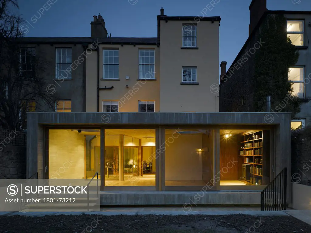 Jigsaw House, Dublin, Ireland. Architect McCullough Mulvin, 2012. View of extension from back garden showing existing house, internal lighting, sliding door closed and american walnut internal door closed at dusk.
