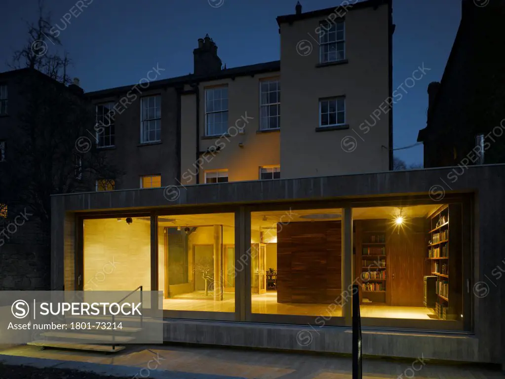 Jigsaw House, Dublin, Ireland. Architect McCullough Mulvin, 2012. View of extension from back garden showing existing house, internal lighting, sliding door closed and american walnut internal door open at dusk.