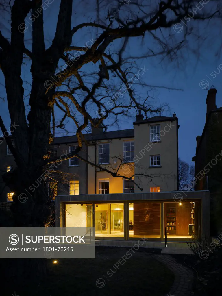 Jigsaw House, Dublin, Ireland. Architect McCullough Mulvin, 2012. View of extension from back garden with tree showing existing house, internal lighting, sliding door closed and american walnut internal door open at dusk.