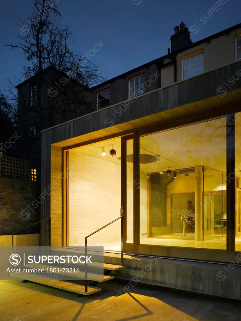 Jigsaw House, Dublin, Ireland. Architect McCullough Mulvin, 2012. View from back garden of sliding door opened showing view to living space and interior lighting at dusk.