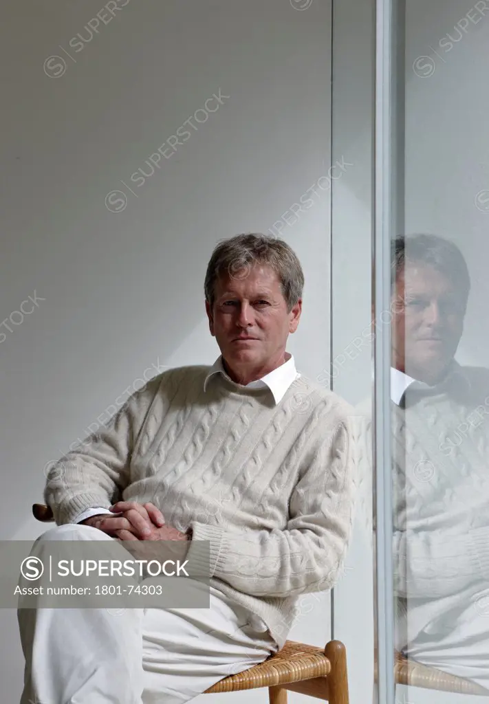 John Pawson at home, London, United Kingdom. Architect: John Pawson, 2010. Portrait of John Pawson sitting on a chair with arms folded in his lap and reflection in glass to his right. Looking into camera.
