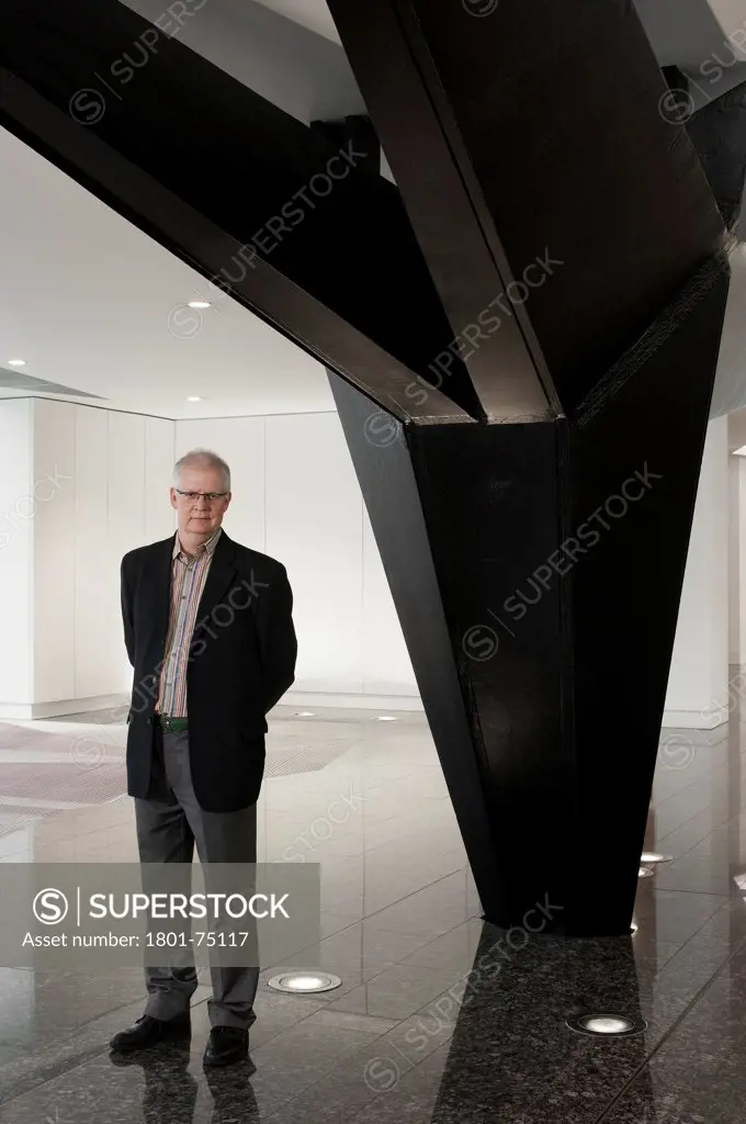 Ken Shuttleworth at 55 Baker Street, London, United Kingdom. Architect: Make Ltd, 2008. General view at reception area looking into lens with neutral expression and arms behind back next to steel supporting column.