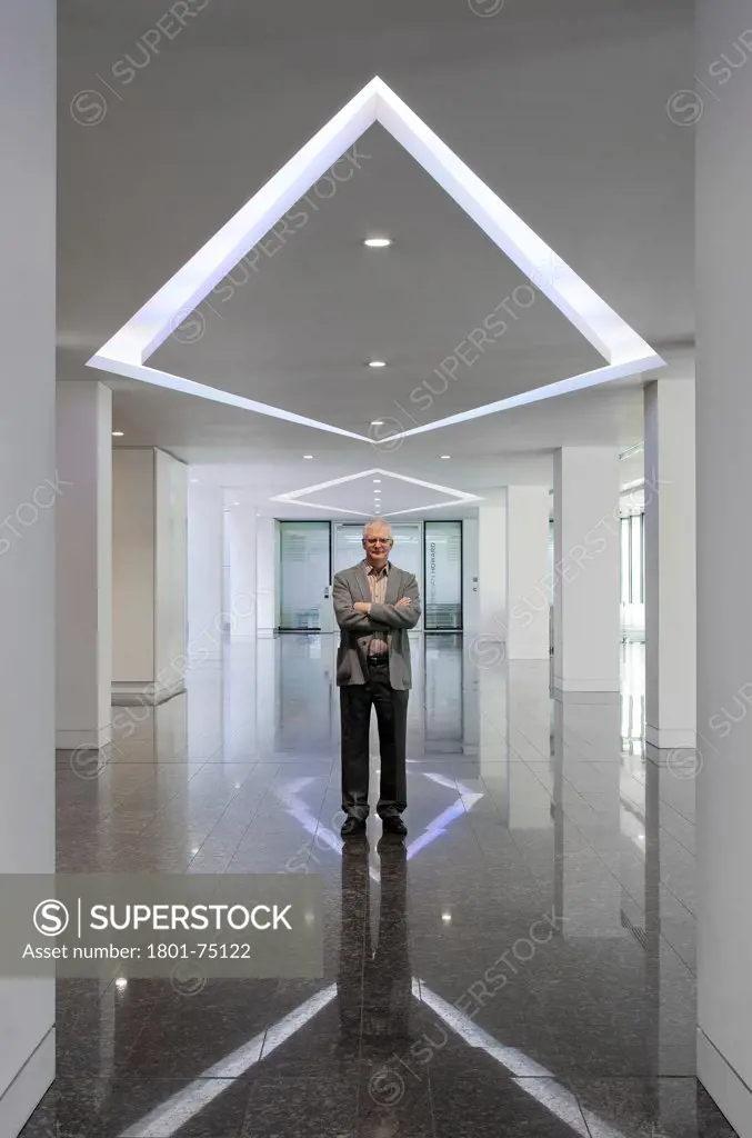 Ken Shuttleworth at 55 Baker Street, London, United Kingdom. Architect: Make Ltd, 2008. Distant view at reception area looking into lens with neutral expression and arms crossed under diamond shaped ceiling lighting.