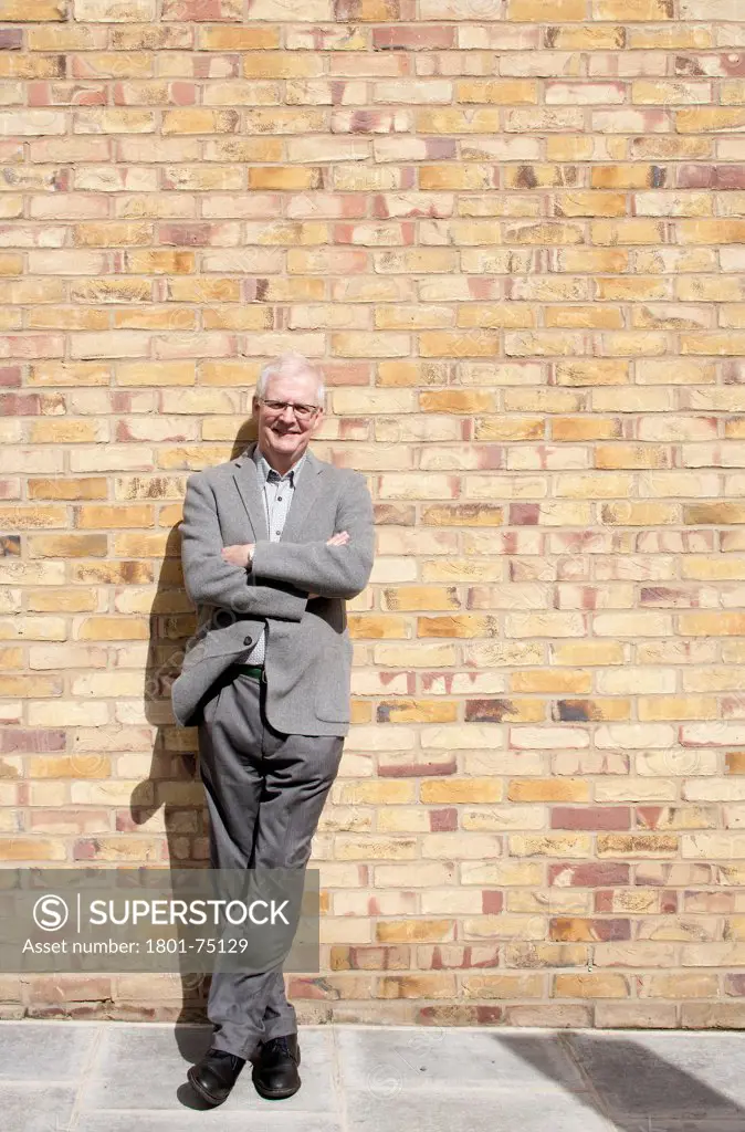 Ken Shuttleworth at 55 Baker Street, London, United Kingdom. Architect: Make Ltd, 2008. General view at residential accomodation looking into lens with smile and arms crossed leaning against brick wall with smile.