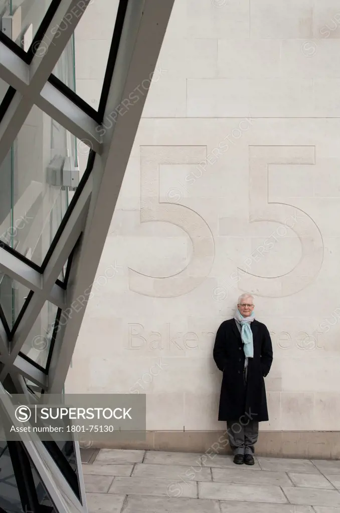 Ken Shuttleworth at 55 Baker Street, London, United Kingdom. Architect: Make Ltd, 2008. Distant view at public entrance looking into lens with smile and arms in pockets leaning against wall with number 55 behind.