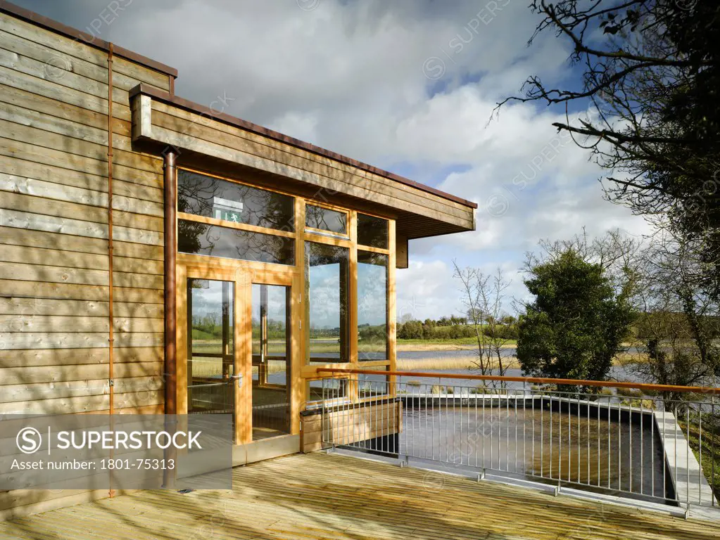 Ballybay Wetland Centre, Ballybay, Ireland. Architect: Solearth Ecological Architecture, 2008. View from decking showing entrance, timber cladding, lake and surrounding landscape.