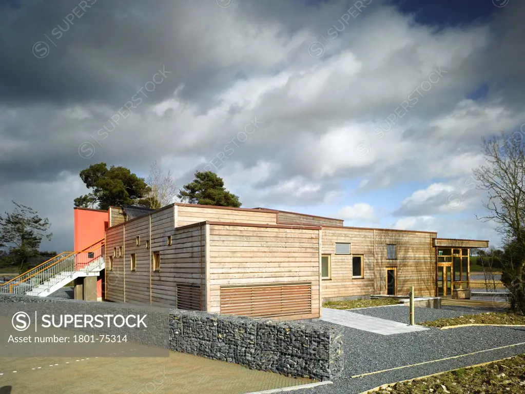Ballybay Wetland Centre, Ballybay, Ireland. Architect: Solearth Ecological Architecture, 2008. View of building showing stone wire mesh wall, timber cladding and surrounding landscape.