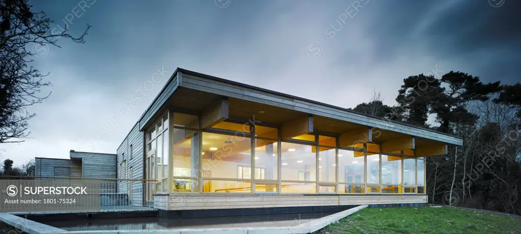 Ballybay Wetland Centre, Ballybay, Ireland. Architect: Solearth Ecological Architecture, 2008. View of building showing timber windows, timber cladding, interior lighting, and surrounding landscape at dusk.