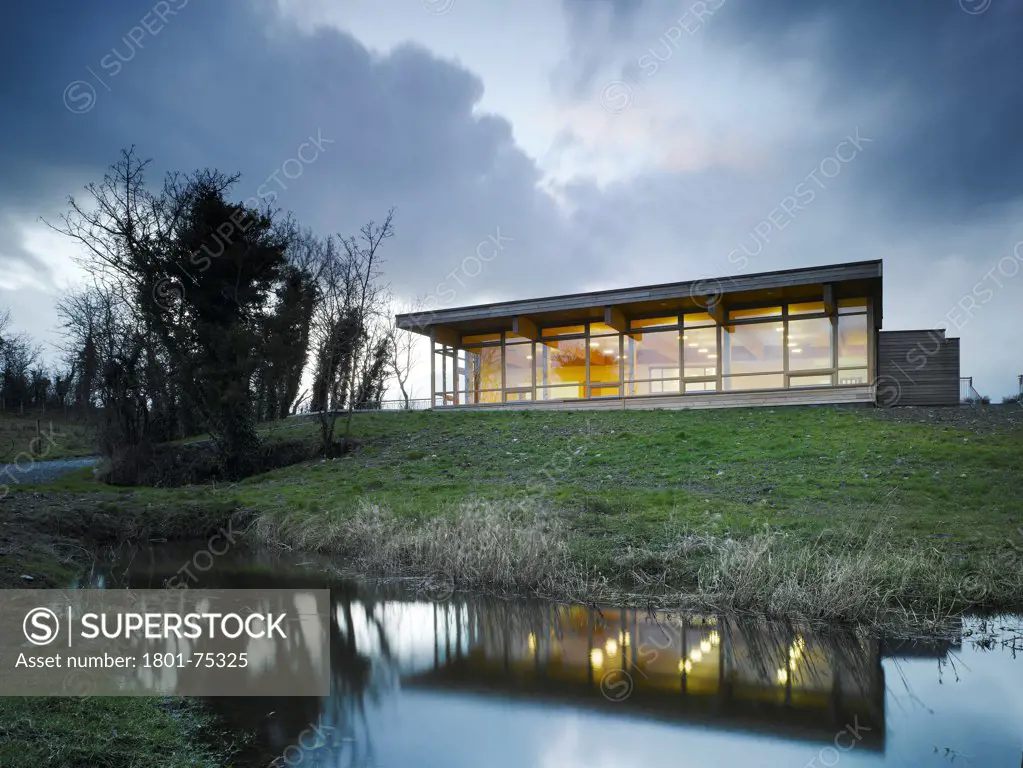 Ballybay Wetland Centre, Ballybay, Ireland. Architect: Solearth Ecological Architecture, 2008. View of building showing timber windows, timber cladding, interior lighting and surrounding landscape at dusk.