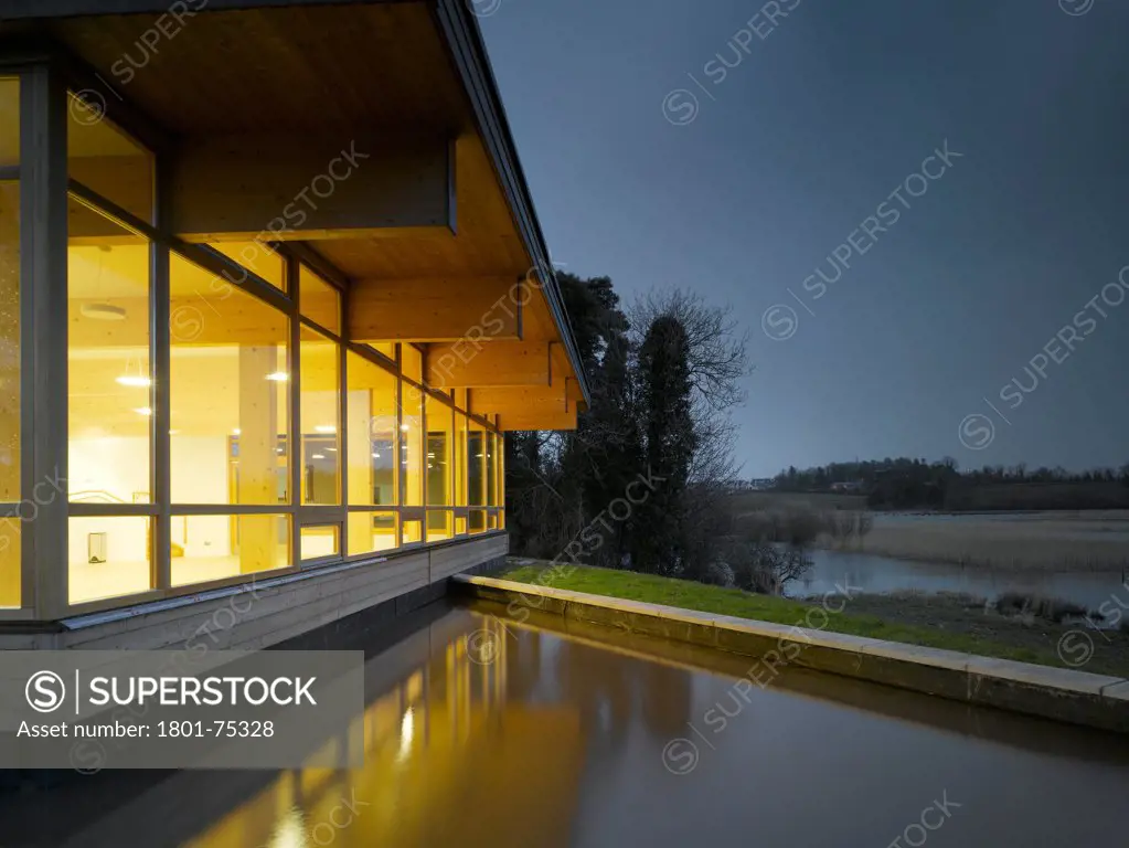 Ballybay Wetland Centre, Ballybay, Ireland. Architect: Solearth Ecological Architecture, 2008. View of building showing surrounding landscape and interior lighting at dusk.