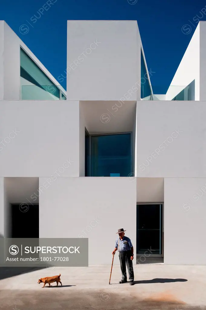 House for Elderly People, Alcaçer do Sal, Portugal. Architect: Francisco Aires Mateus Arquitectos, 2011. View of social housing complex in daylight with man in hat and dog.