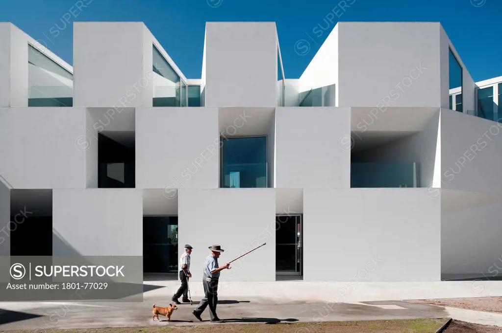 House for Elderly People, Alcaçer do Sal, Portugal. Architect: Francisco Aires Mateus Arquitectos, 2011. View of social housing complex in daylight with man in hat and dog.