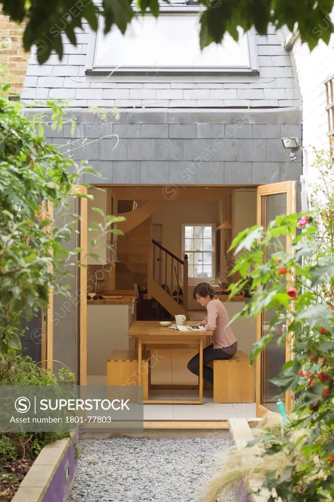 The Slim House, Terraced House, Europe, United Kingdom, , 2012, alma-nac architects. Detail rear view of house with doors open and woman reading at table.