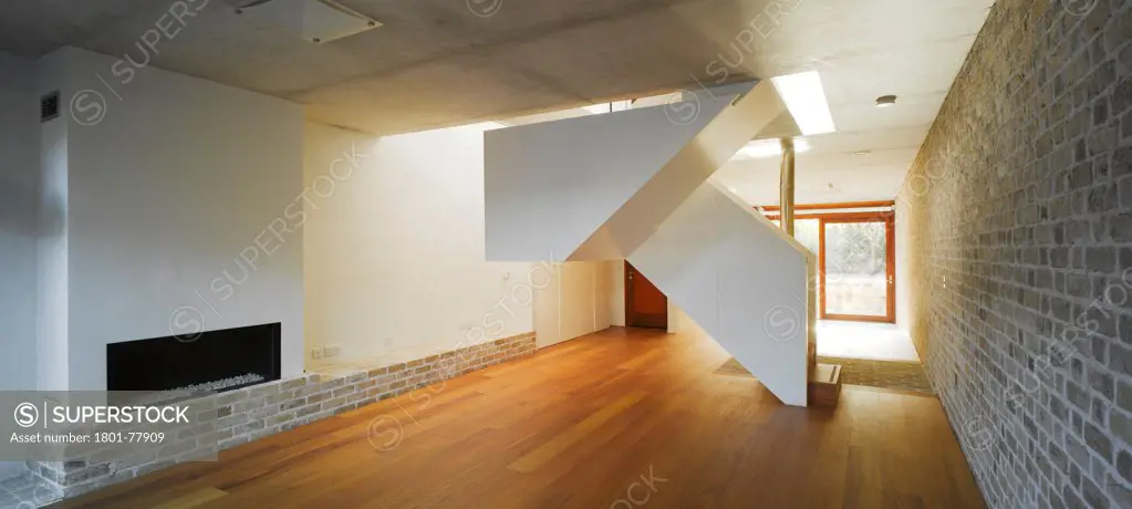 Mews Houses, Dublin, Ireland. Architect: Grafton Architects, 2009. View of main living space showing fireplace, stairs, view to exterior, exposed brick wall and timber flooring.