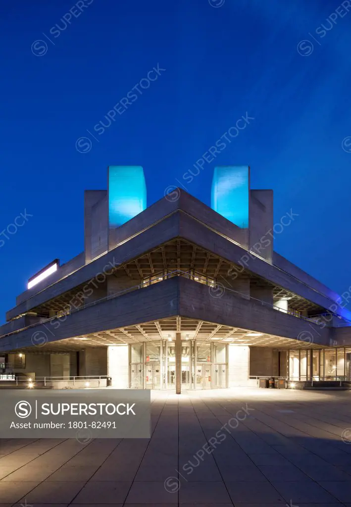 Royal National Theatre, London, United Kingdom. Architect: Denys Lasdun, 1975. Night shot of the main foyer entrance to the theatre with light spilling from the interior and a dramatic sky.