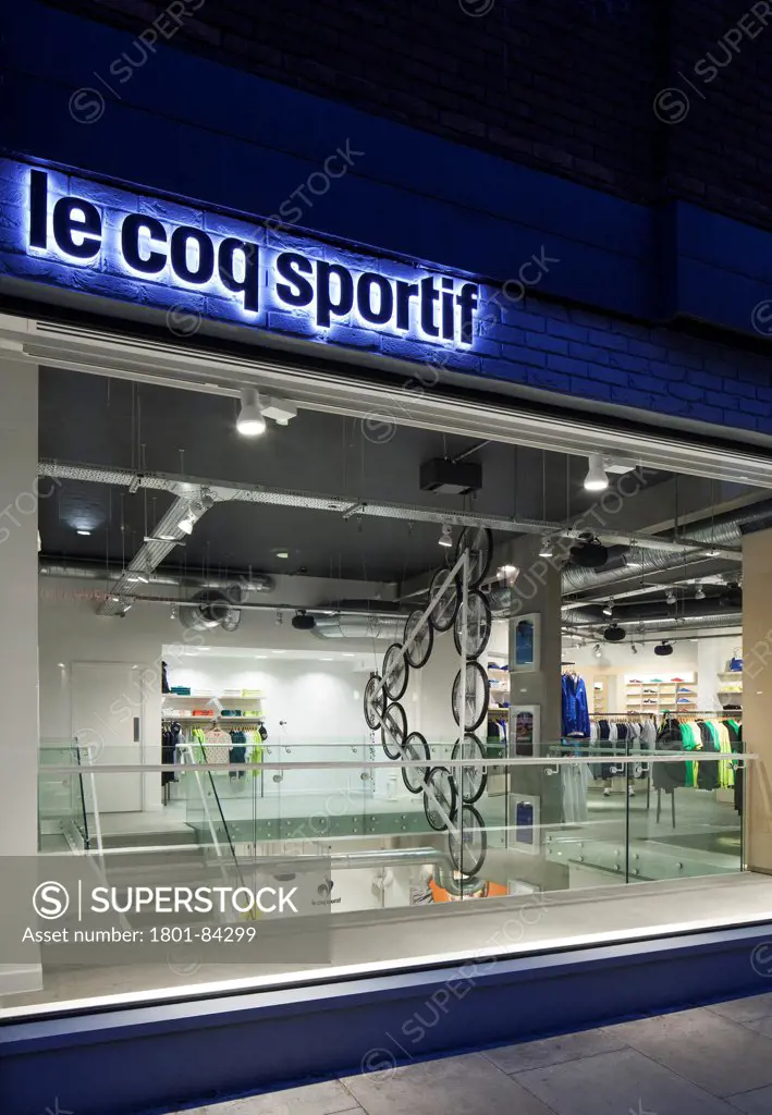 Le Coq Sportif London -FlagshipStore, London, United Kingdom. Architect Studio Oscar, 2013. View looking into the store at night with branding and signage,Kinetic bicycle sculpture can be hangs over the stairs.