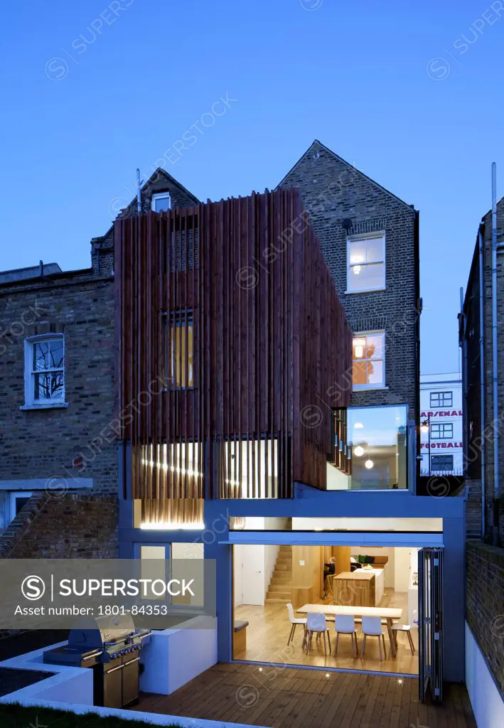Power House, London, United Kingdom. Architect Paul Archer Design, 2013. View from the garden showing thesculptural timber clad rear extension at dusk. The old Arsenal stadium can be seen through the gap between the buildings.