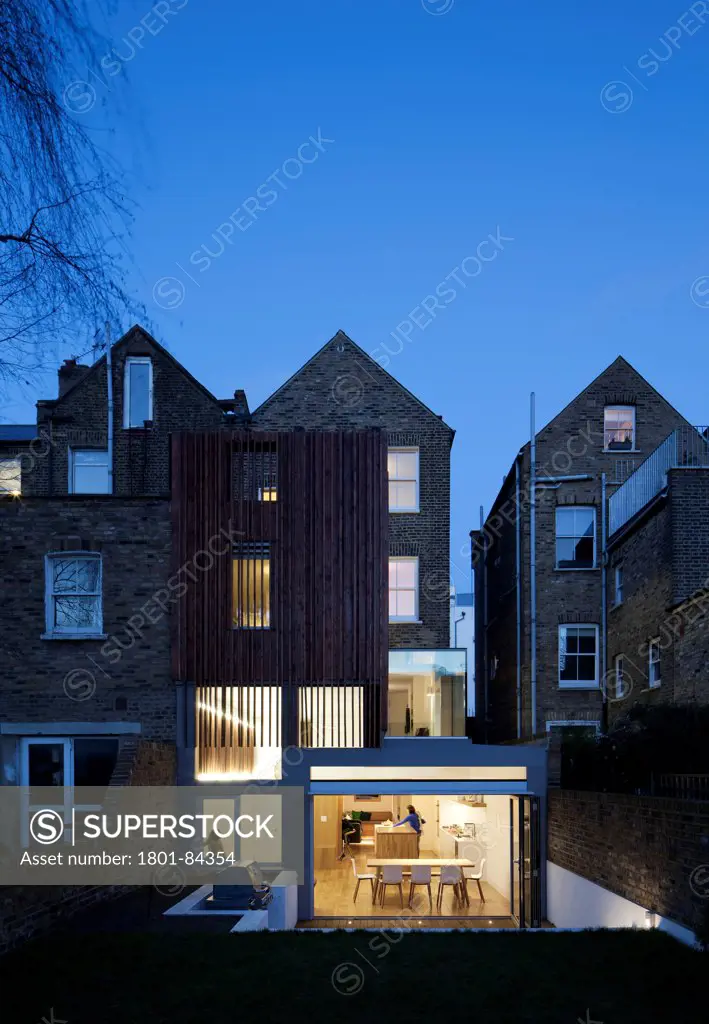 Power House, London, United Kingdom. Architect Paul Archer Design, 2013. View from the garden showing sculptural timber clad rear extension at dusk. The occupants can be seen through the window.