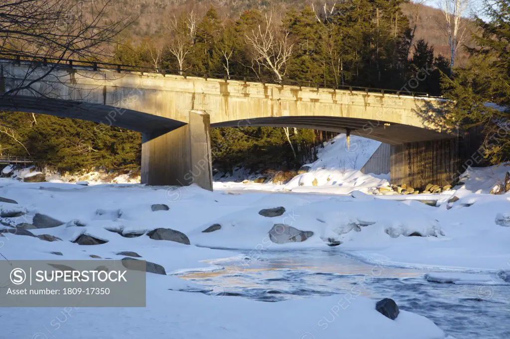 Road Bridge during the in winter months. This bridge crosses the East Branch of the Pemigewasset River in Lincoln, New Hampshire USA along Kancamagus Scenic Byway (Route 112). A suspension bridge used for foot traffic is in the background and a nearly ful