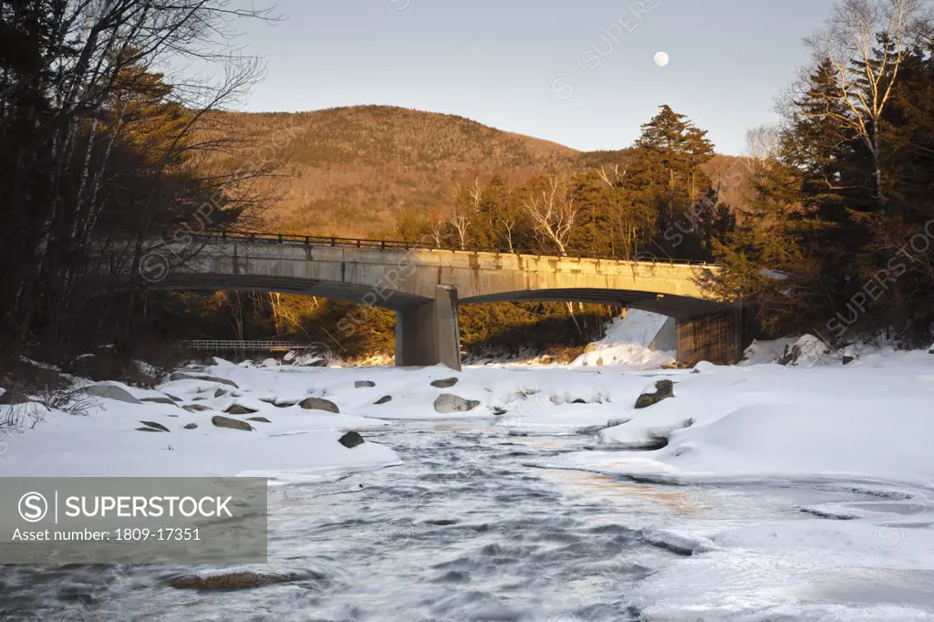 Road Bridge during the in winter months. This bridge crosses the East Branch of the Pemigewasset River in Lincoln, New Hampshire USA along Kancamagus Scenic Byway (Route 112). A suspension bridge used for foot traffic is in the background and a nearly ful