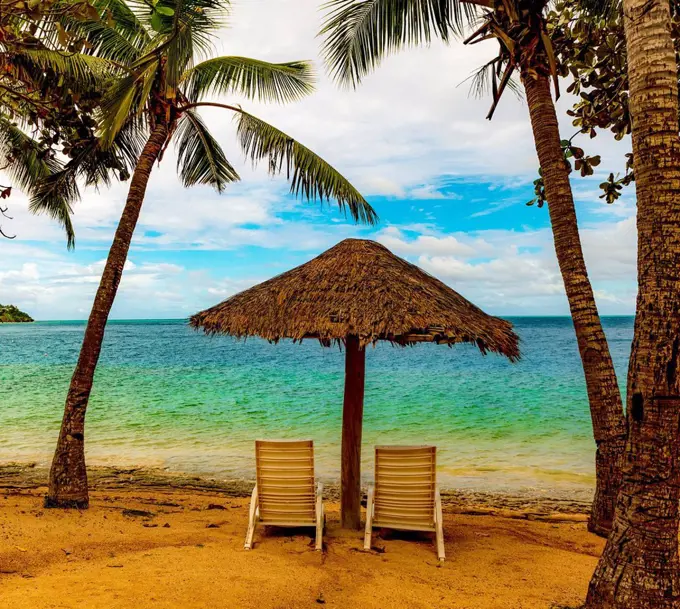 Two chairs under a thatched umbrella on the beach looking out to the turquoise ocean water; Malolo Island, Fiji
