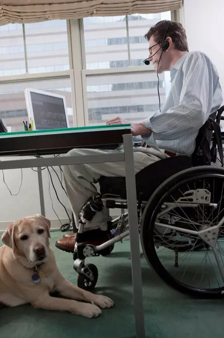 Man in a wheelchair with a Spinal Cord Injury and a headset working at desk in a home office with a service dog