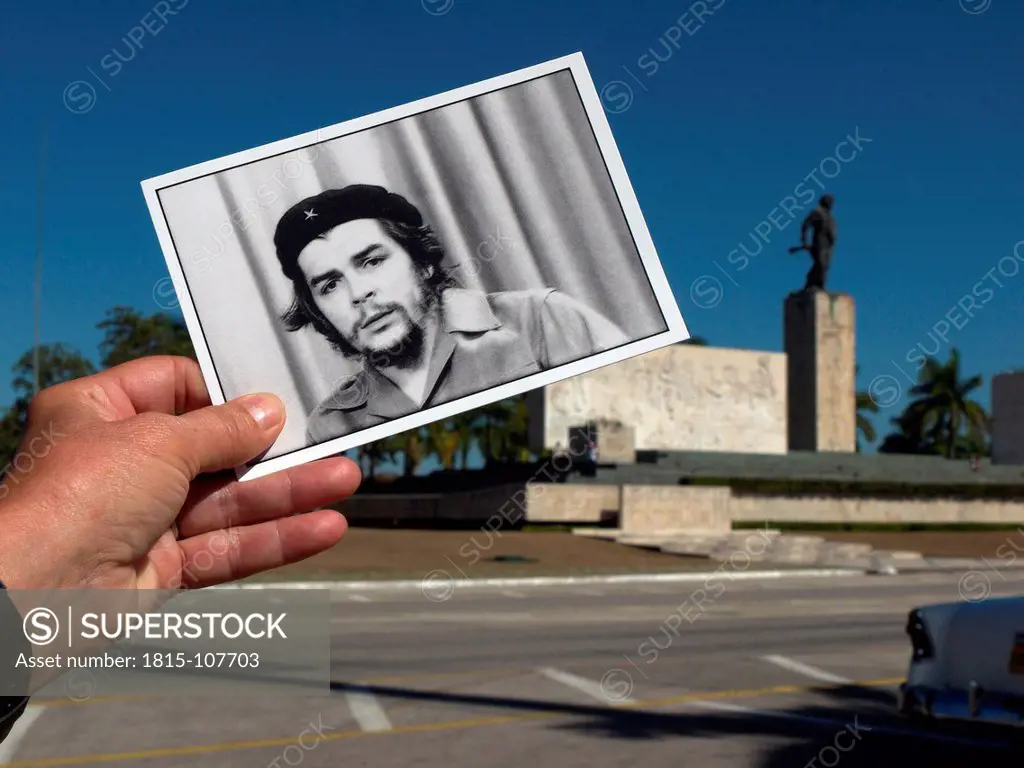 Cuba, Santa Clara, Close up of hand holding Che Guevara postcard with Memorial del Ernesto Che Guevara in background