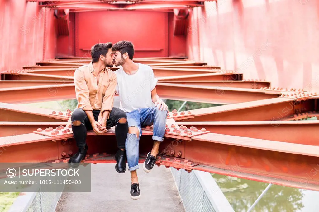 Kissing young gay couple sitting on steel girder of a footbridge