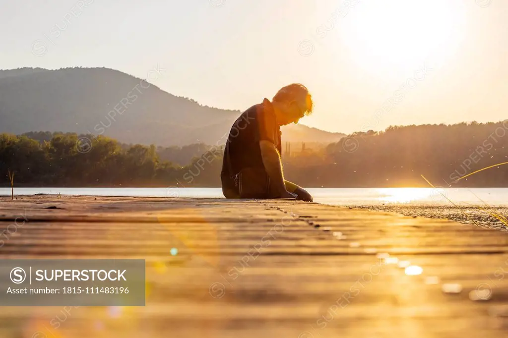 Senior man sitting on a wooden footbridge and looking down, against the sun