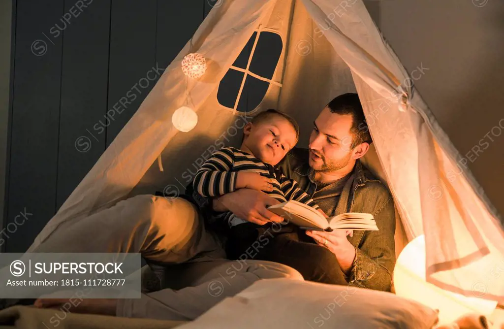 Father reading book to son at an illuminated tent at home
