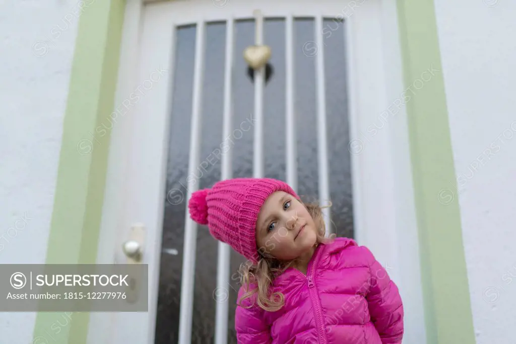 Young girl wearing pink winter jacket and cap, standing in front of entry door