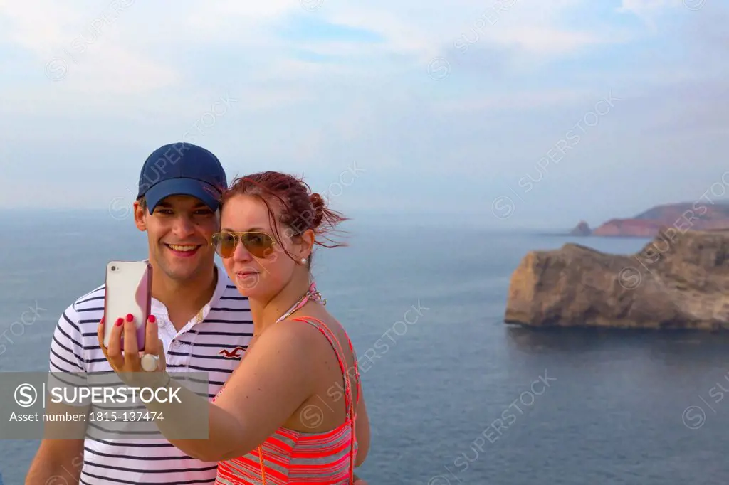 Portugal, Couple taking photo with iphone at Cape Sao Vicente, smiling