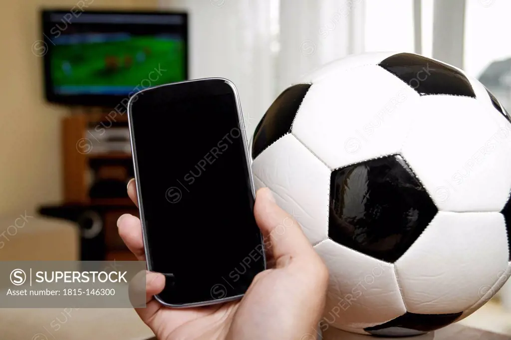 Mature man holding mobile with soccer ball and TV in background