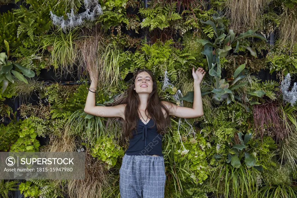 Portrait of smiling young woman with closed eyes in front of plant wall
