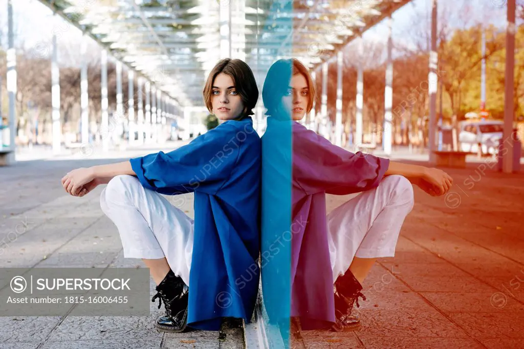 Young woman with urban look sitting on the floor and leaning on colorful glass wall with her reflection