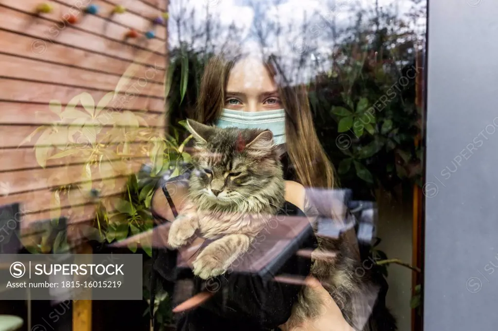 Portrait of girl with surgical mask and cat behind window pane