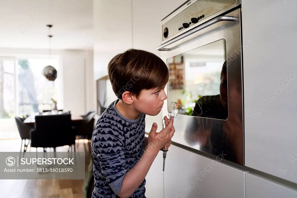 Boy tasting batter while watching his mirror image on surface of oven