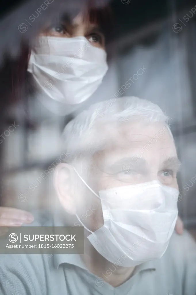 Portrait of senior couple wearing masks at home, looking out of the window