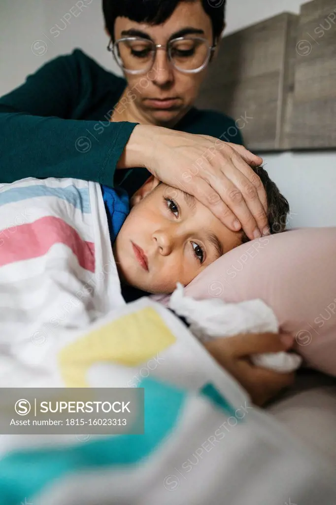 Portrait of sick boy lying in bed while his mother touching his forehead
