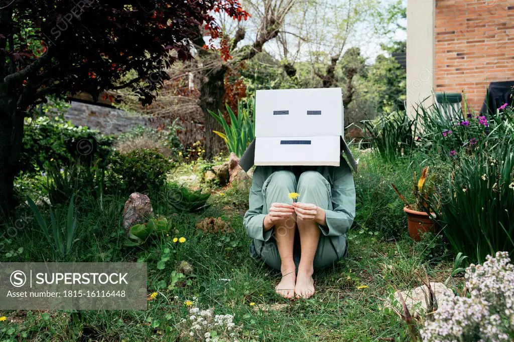 Young woman with cardboard box on her head sitting barefoot in garden