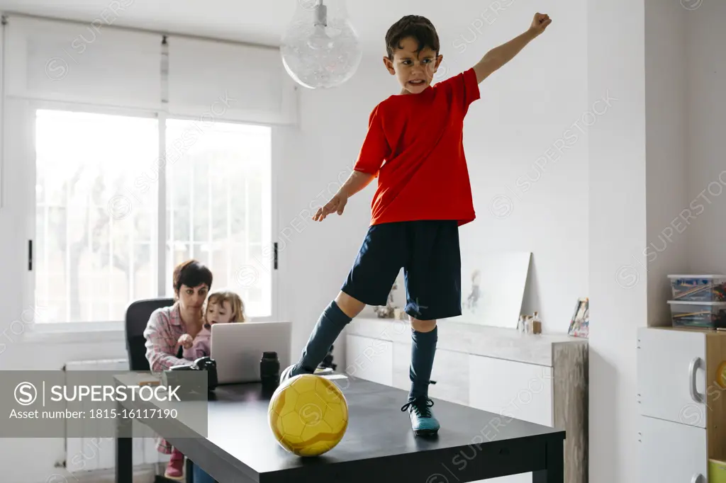 Mom working at home with her children, son standing on soccer ball on table raising arm