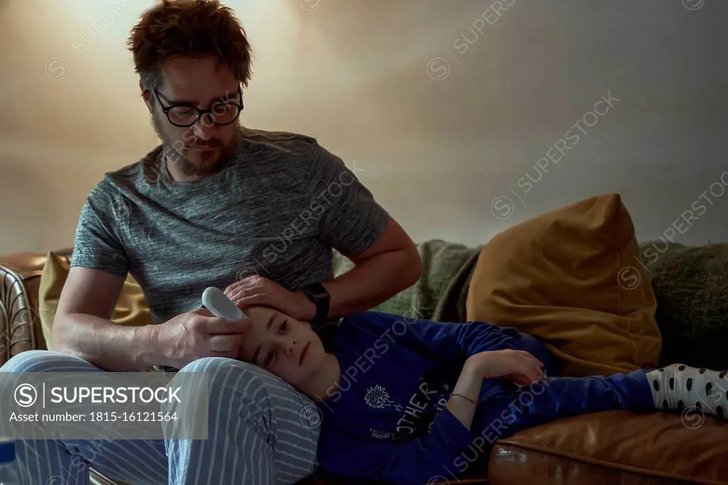 Man holding thermometer on sick daughter's forehead in living room at home