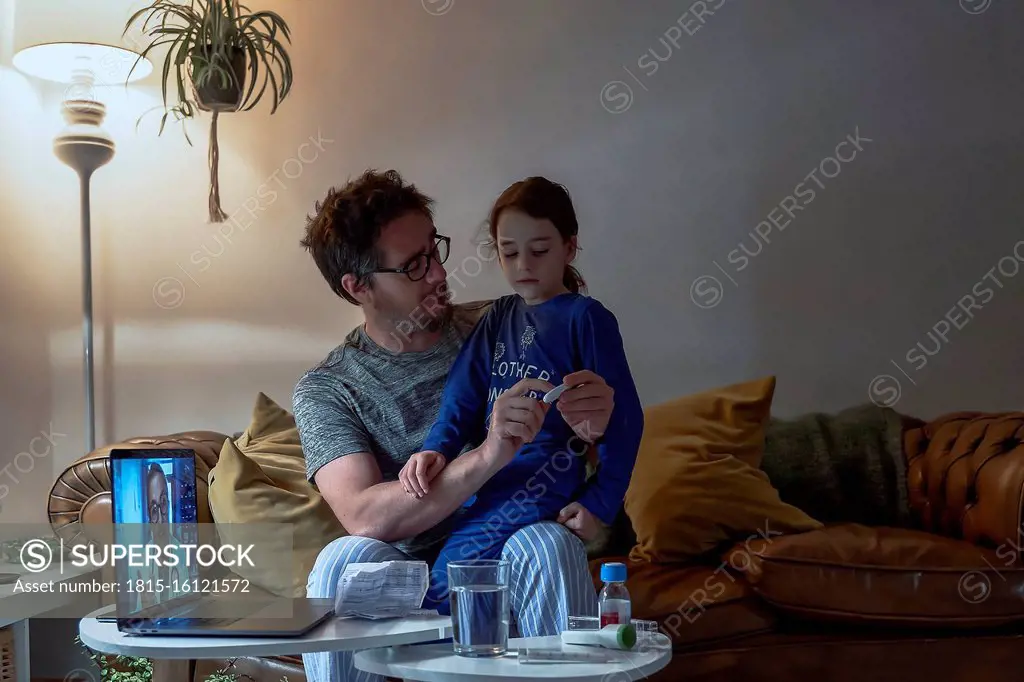 Man holding thermometer while sitting with sick daughter during video call at home