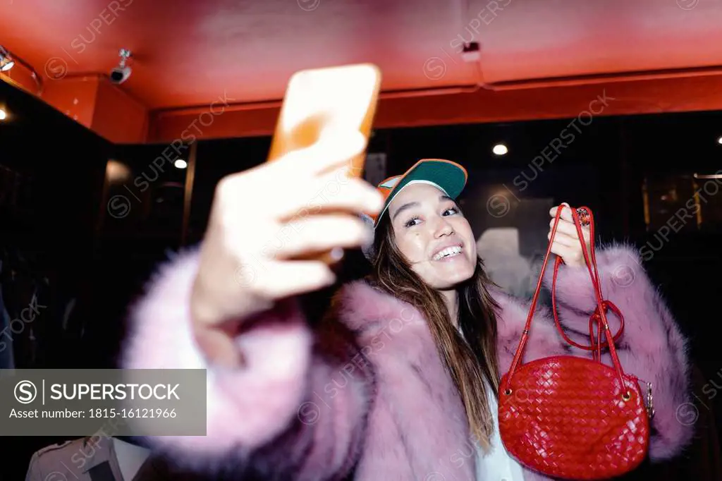 Happy woman taking selfie through smart phone while wearing fur jacket and holding leather purse in store